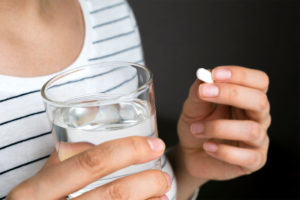 Close up of woman holding a white oval pill and a glass of water.