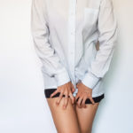 A close-up image of a woman in a white button-up shirt and black shorts gripping her bare thighs with her fingers as her legs are crossed.