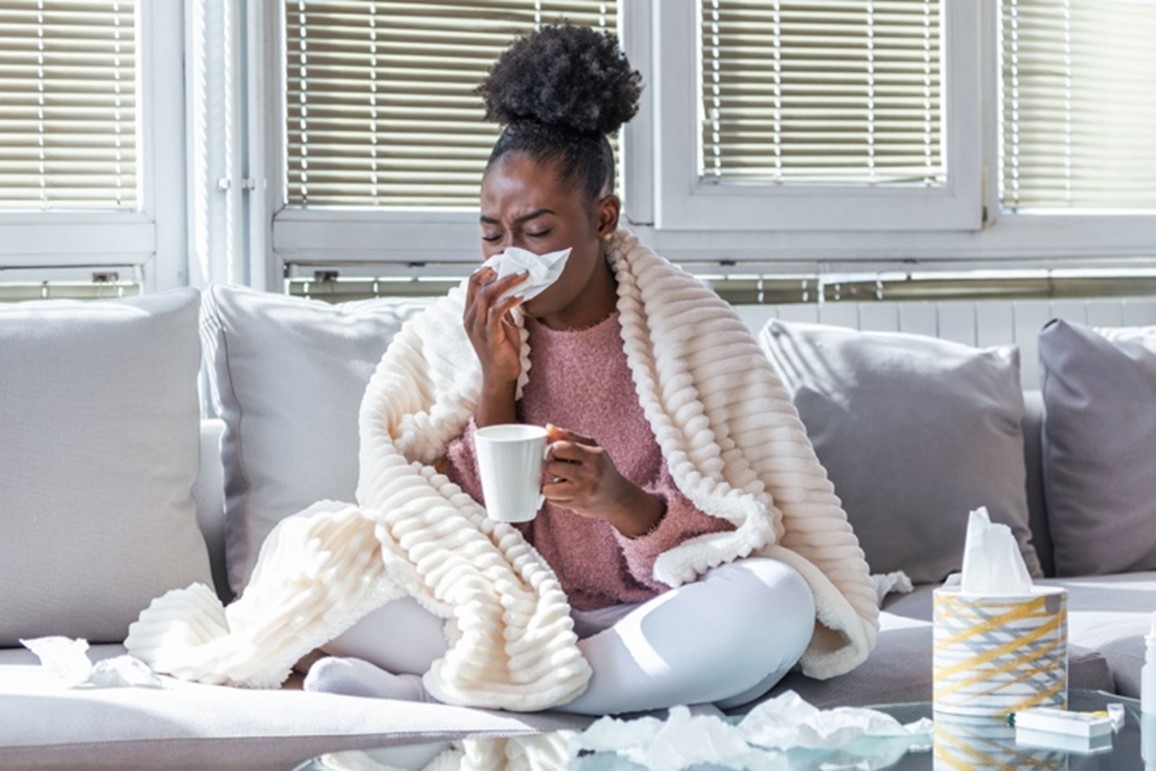A sick woman sits on her sofa wrapped in a blanket, holding a coffee cup in one hand and a tissue to her nose in the other.