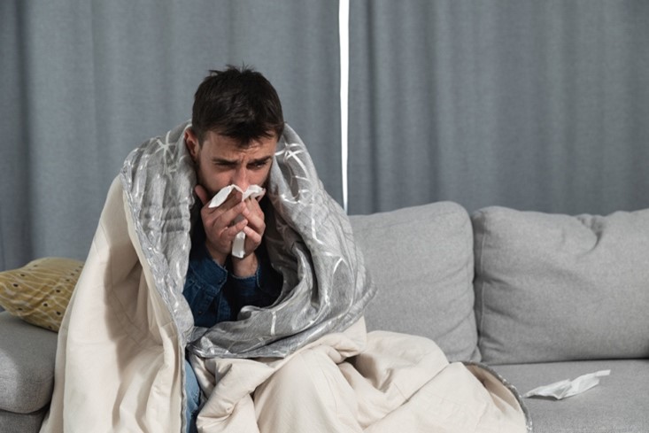 A sick man sits on a couch wrapped in a blanket blowing his nose.