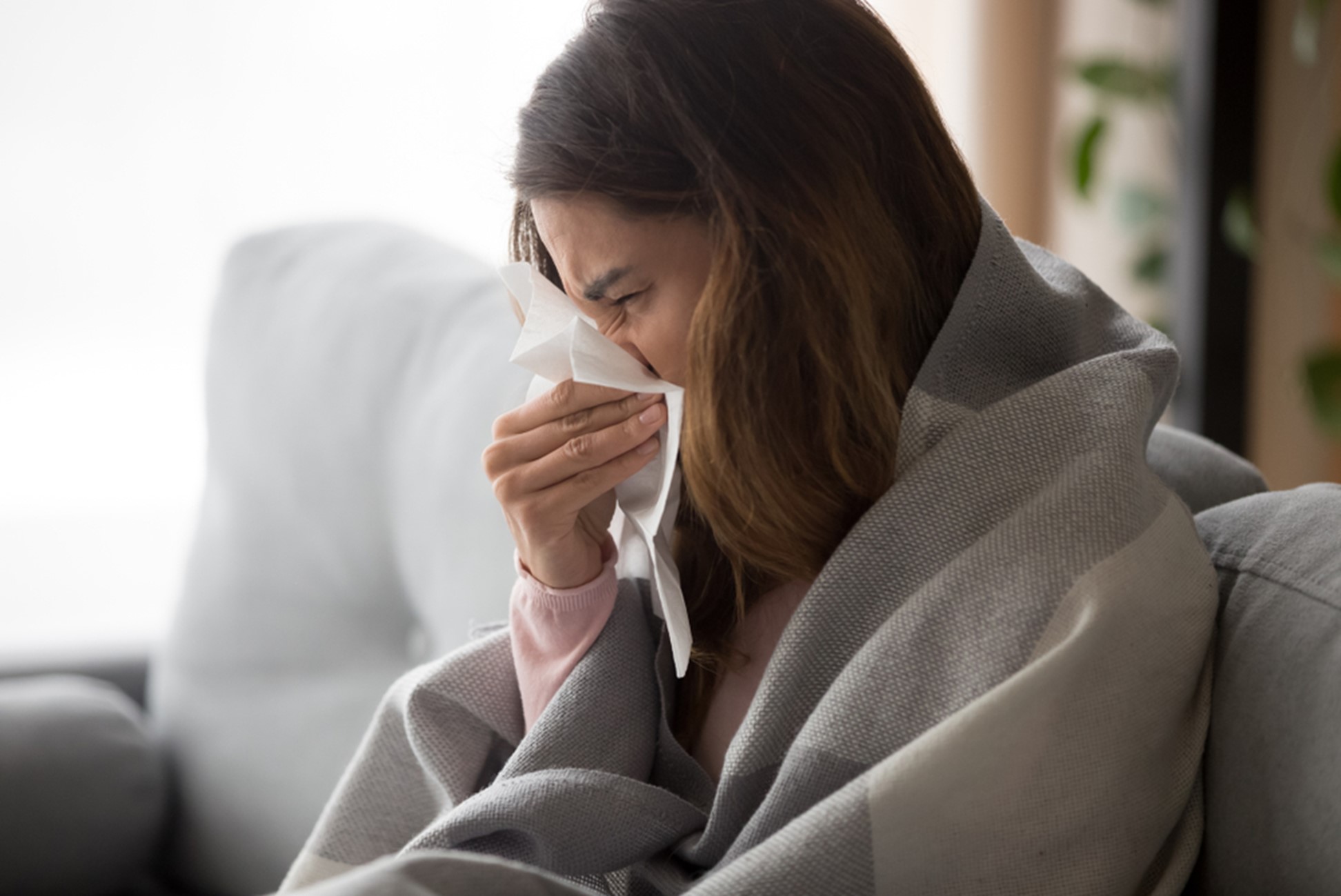 A closeup of a woman sitting on a sofa wrapped in a blanket and blowing her nose into a tissue.