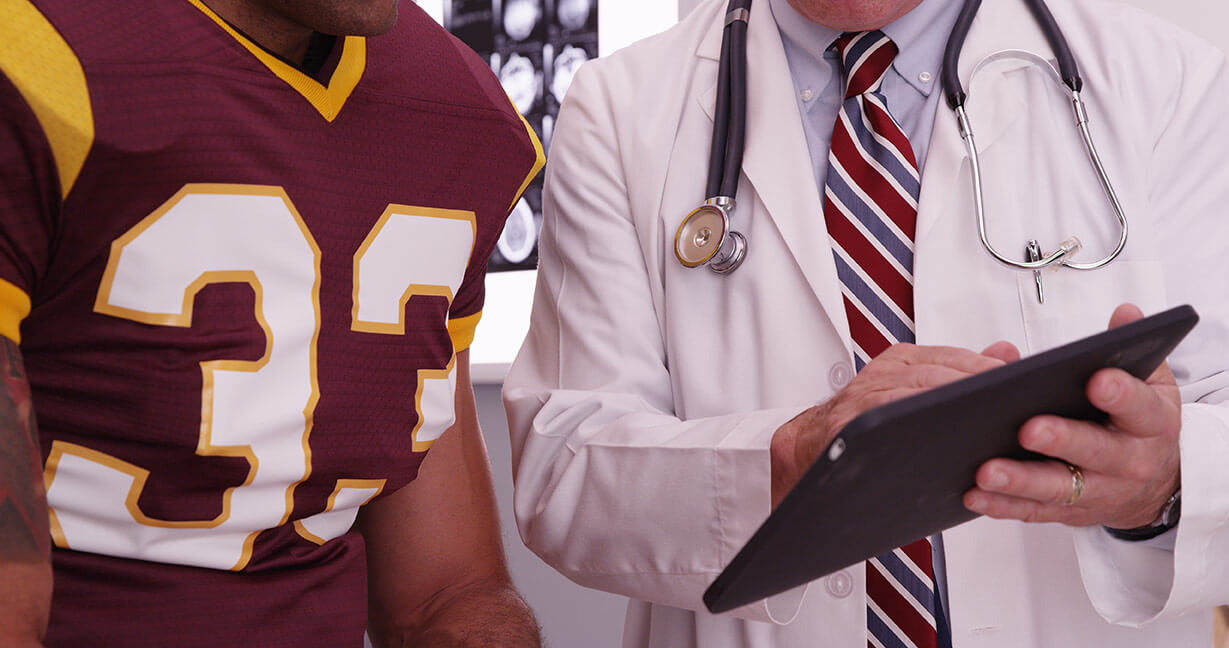 A closeup of a medical doctor reviewing a patient chart with a patient wearing a football jersey.