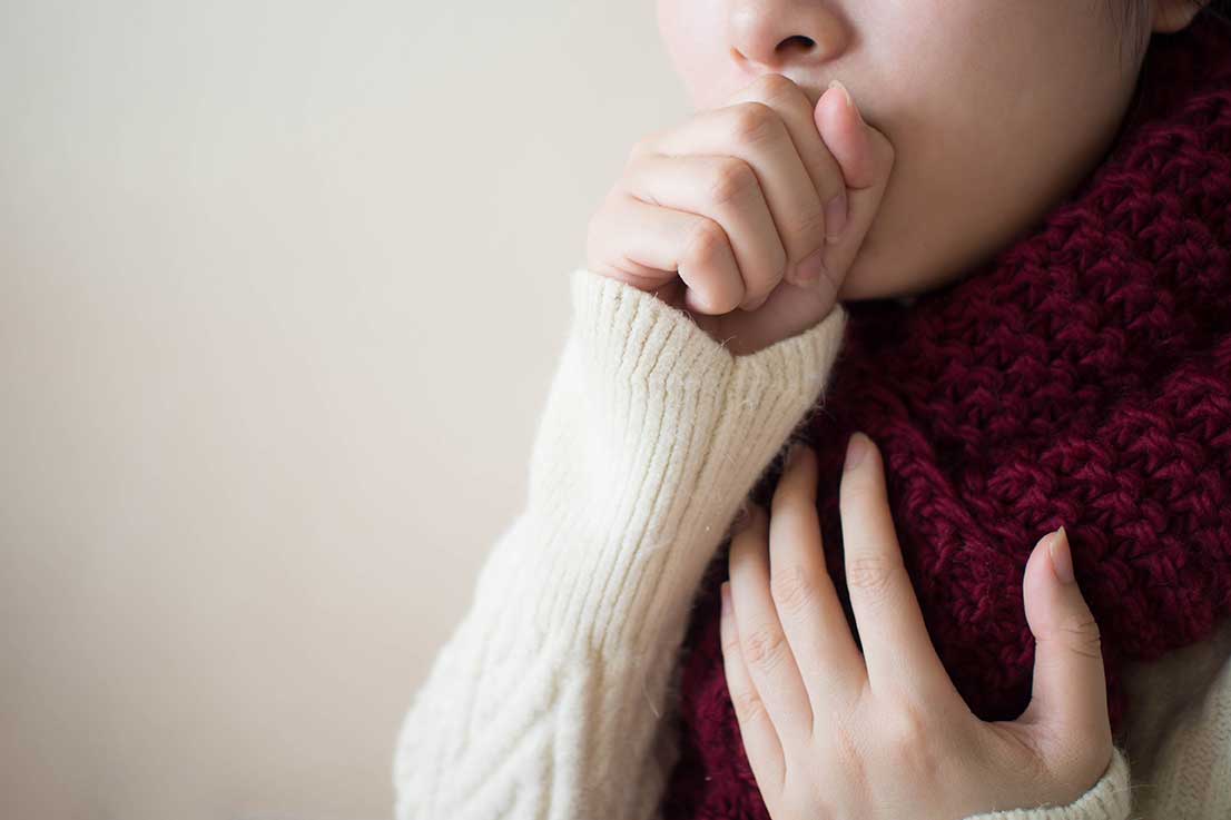 A close-up image of a female with a sweater and a scarf on holding her chest and coughing into a clenched fist.