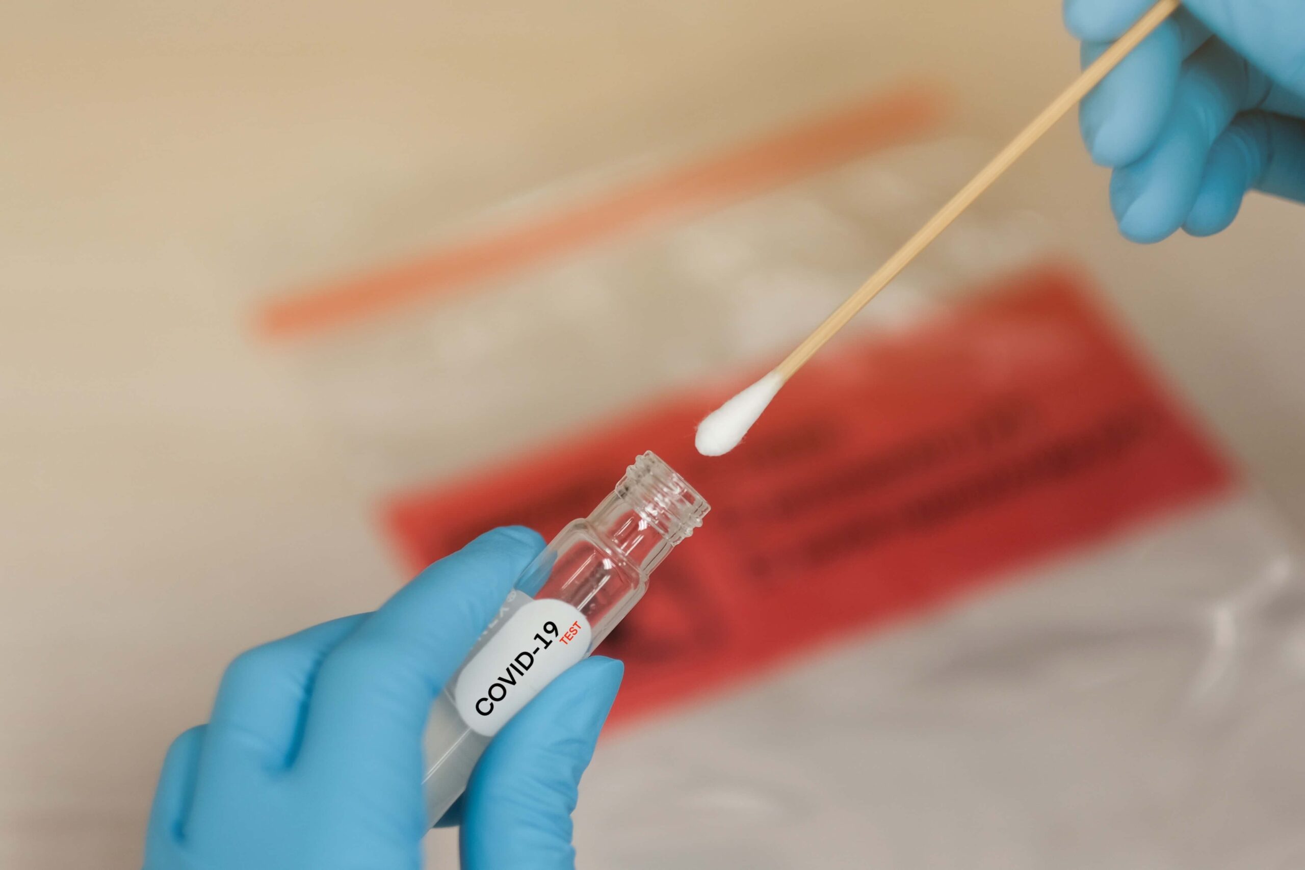 Blue-gloved hands placing a nasal swab into a laboratory vial labeled “COVID-19 with a biohazard bag blurred in the background.