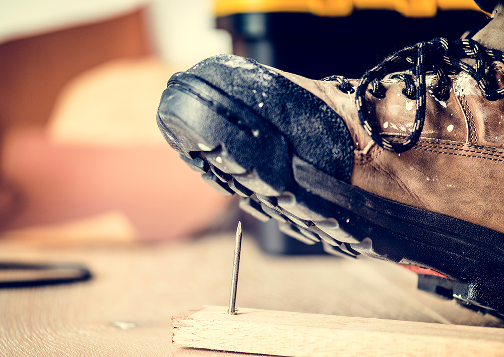 A closeup image of a work boot hovering over a nail in a piece of wood.