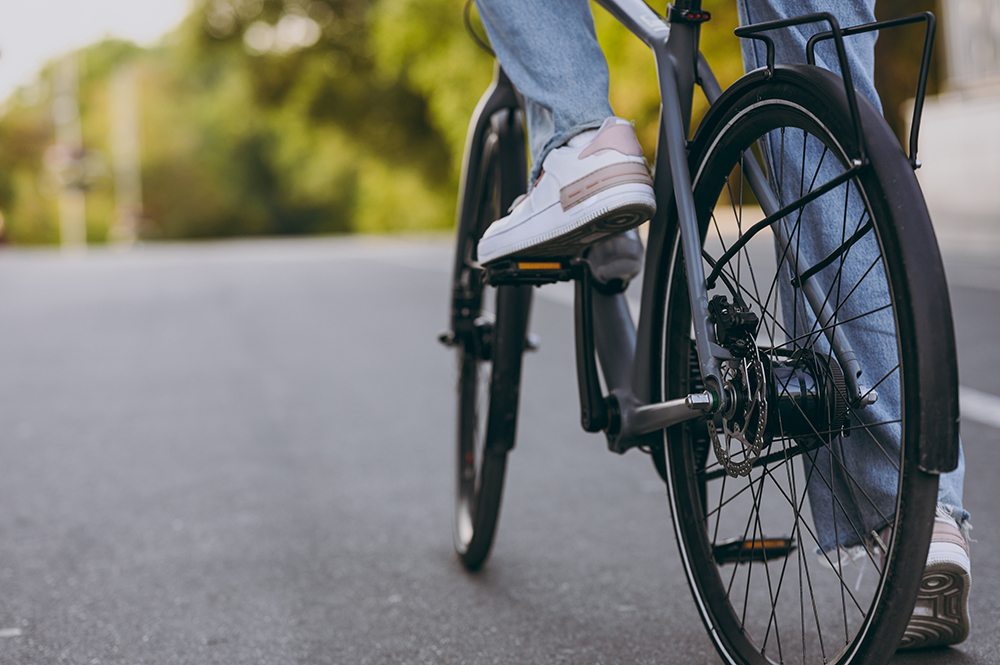 A close-up view of a person riding a bicycle on a paved road, showing the rear wheel, gear system, and sneakers pedaling.