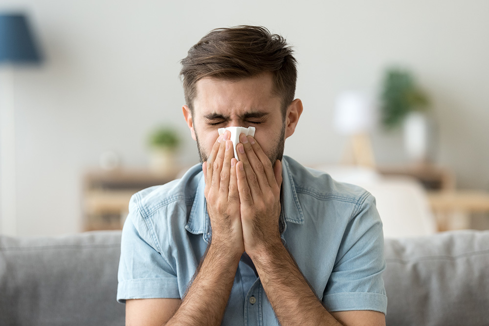 A man sits on a couch with his eyes closed, holding a tissue to his nose with both hands.